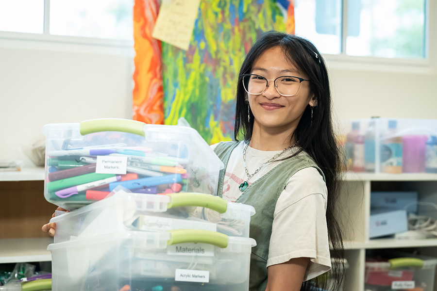 Teresa Lam, smiling in glasses and a sage green vest, carries stacked clear bins of labeled art supplies in front of a colorful painting.