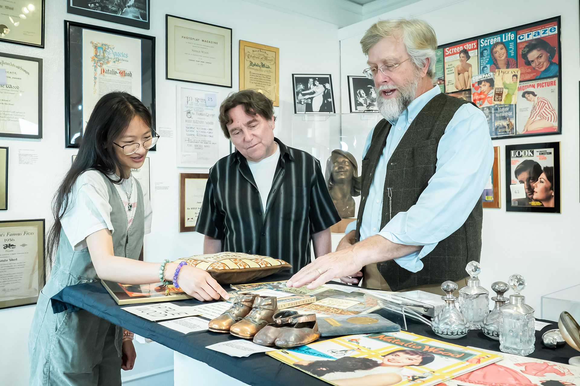 Teresa with two men reviewing what looks like Natalie Wood memorabilia — bronzed baby shoes, vintage magazines, cut-out doll books — in an exhibit or archive room. 