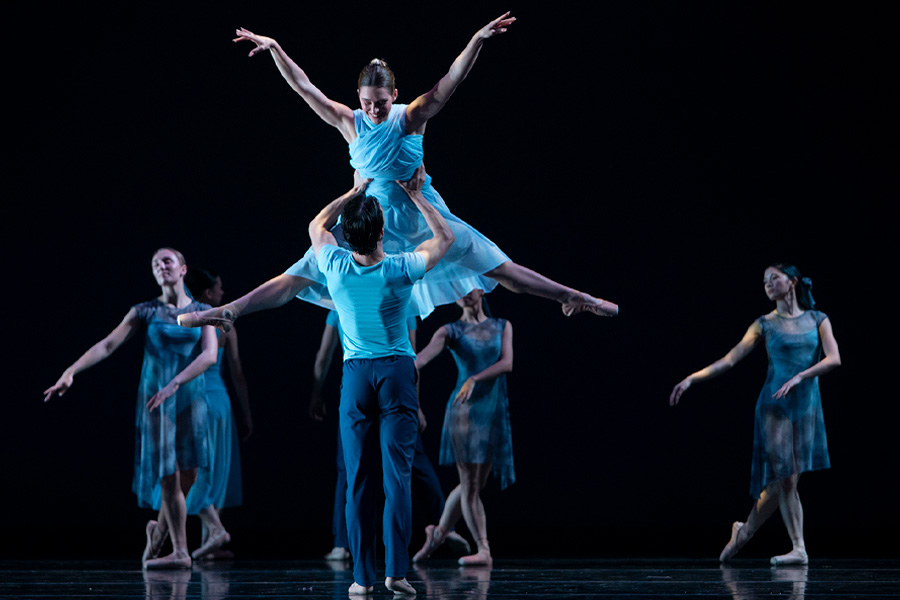 A male dancer lifts a female dancer overhead in a dramatic pose, her arms outstretched, as an ensemble of dancers in flowing blue dresses moves across a darkened stage.