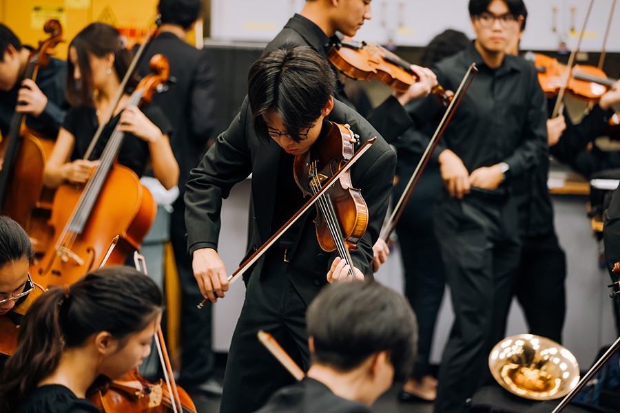 UCI Symphony Orchestra students tune their instruments backstage before a performance, with violinists, cellists, and a French horn player visible.