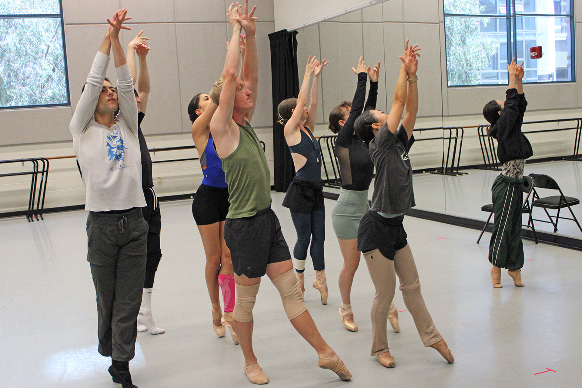 Dance students in a studio reach their arms overhead in unison during a ballet class, wearing rehearsal clothes and pointe shoes, with mirrors and barres lining the walls behind them.