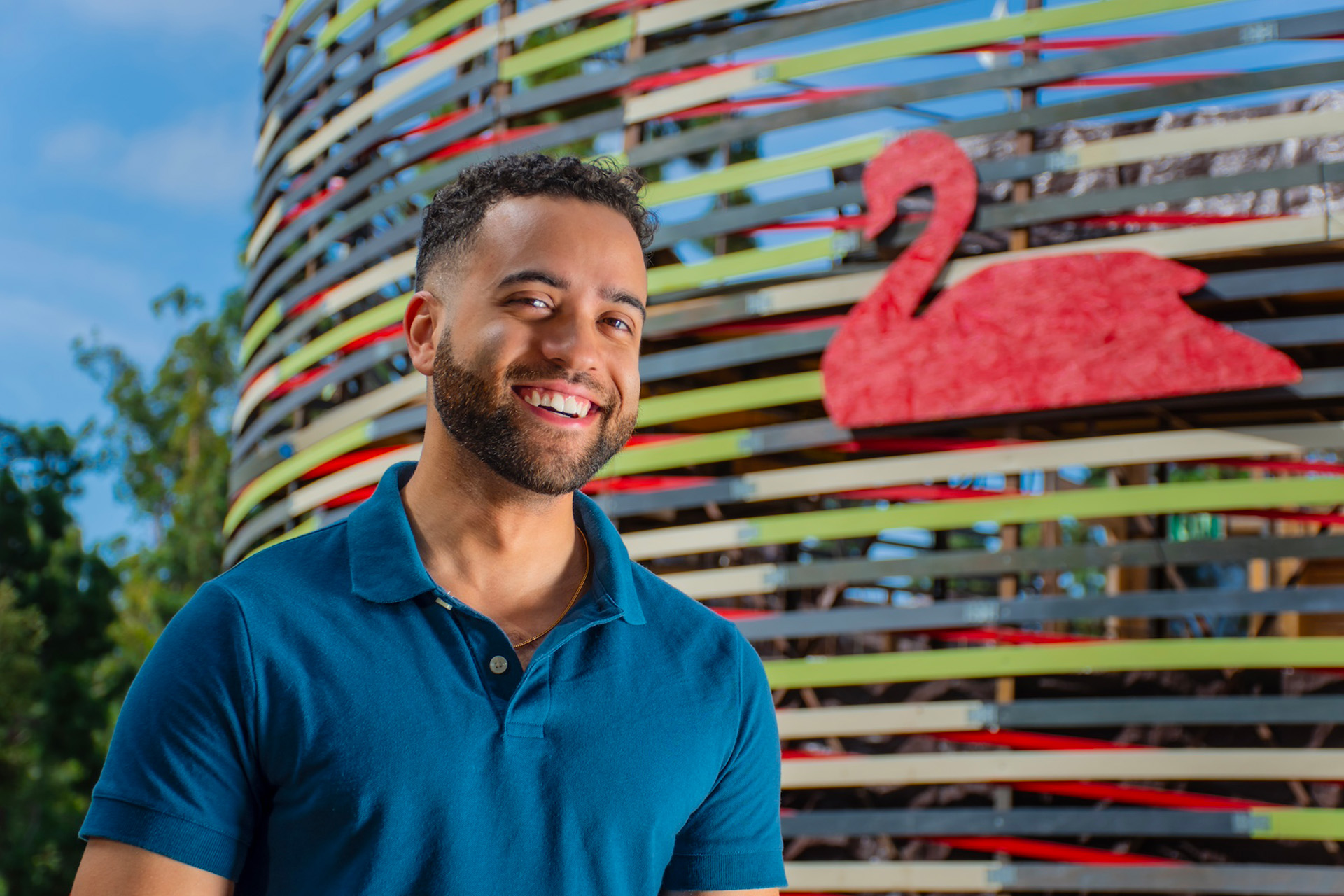 Evan Lugo, wearing a teal polo shirt, smiles while standing in front of the New Swan Theater's curved slat exterior, which features a stylized red swan design, on a sunny day.