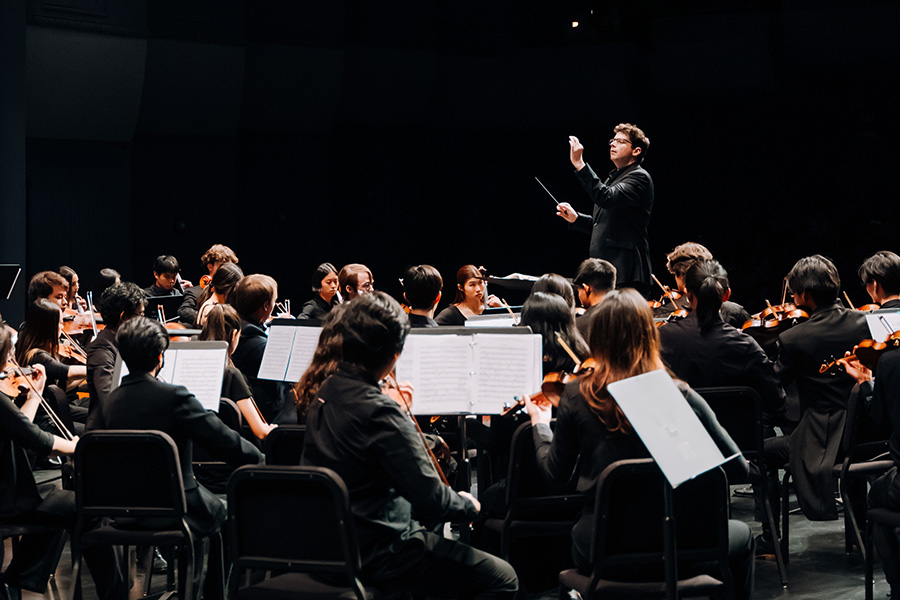 Geoffrey Pope conducts the full UCI Symphony Orchestra on stage, viewed from behind the ensemble with musicians and sheet music in the foreground.
