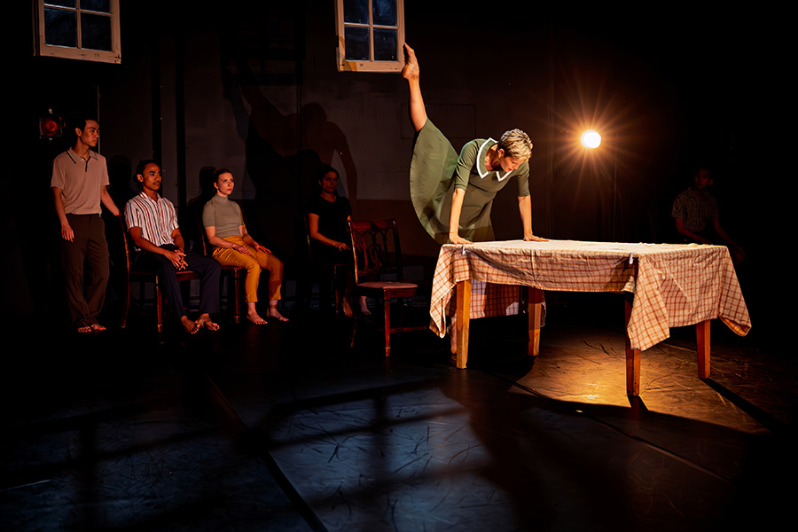 Boróka Nagy, in a green dress, balances on a table with her leg extended overhead as seated cast members look on.