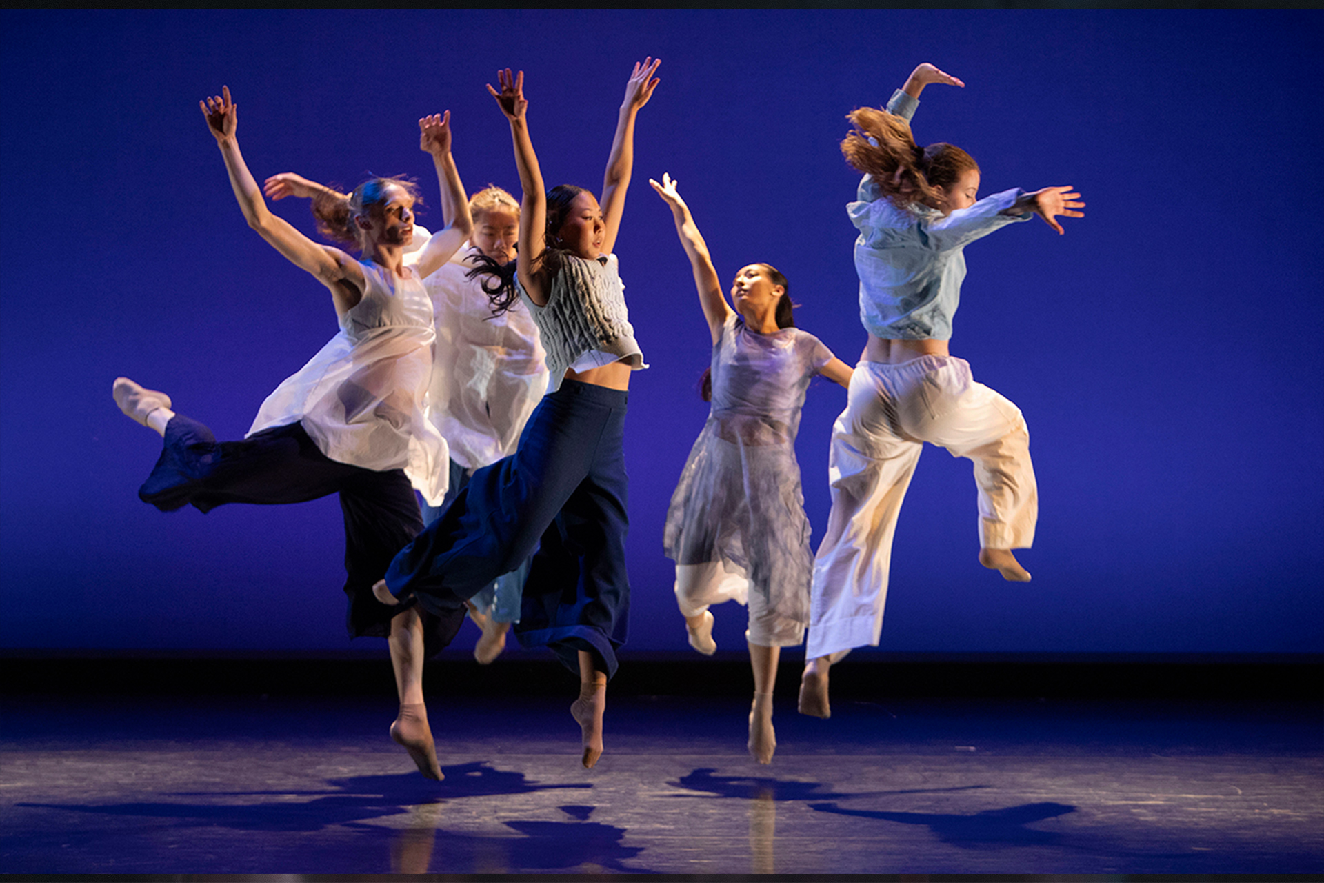 Five dancers leap simultaneously on a dimly lit stage, arms raised, wearing flowing white and navy garments against a deep blue backdrop.