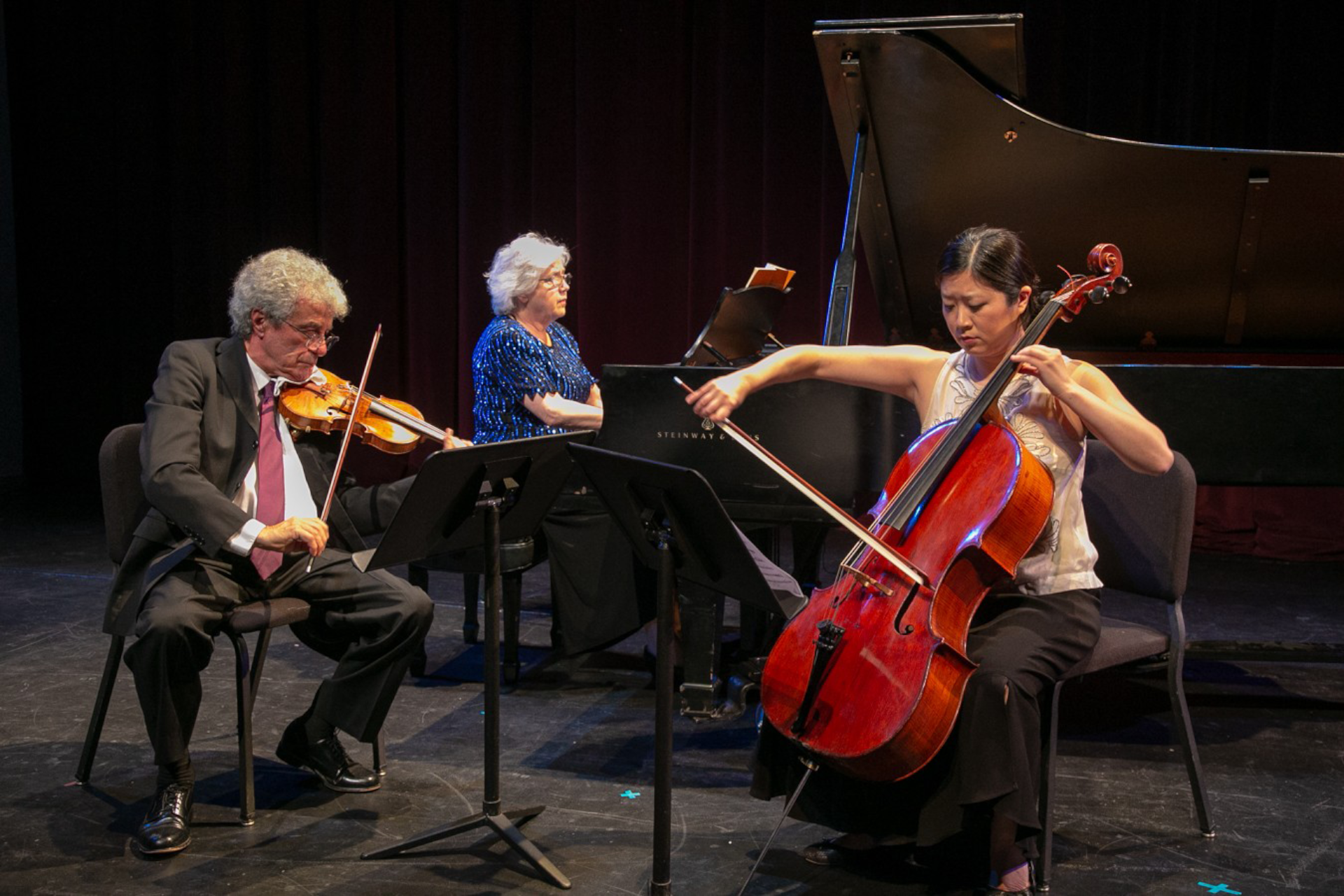 Faculty musicians perform on a darkened stage, with Haroutune Bedelian playing violin on the left, Lorna Griffitt at a grand piano in the center, and Sarah Koo Freeman playing cello on the right.