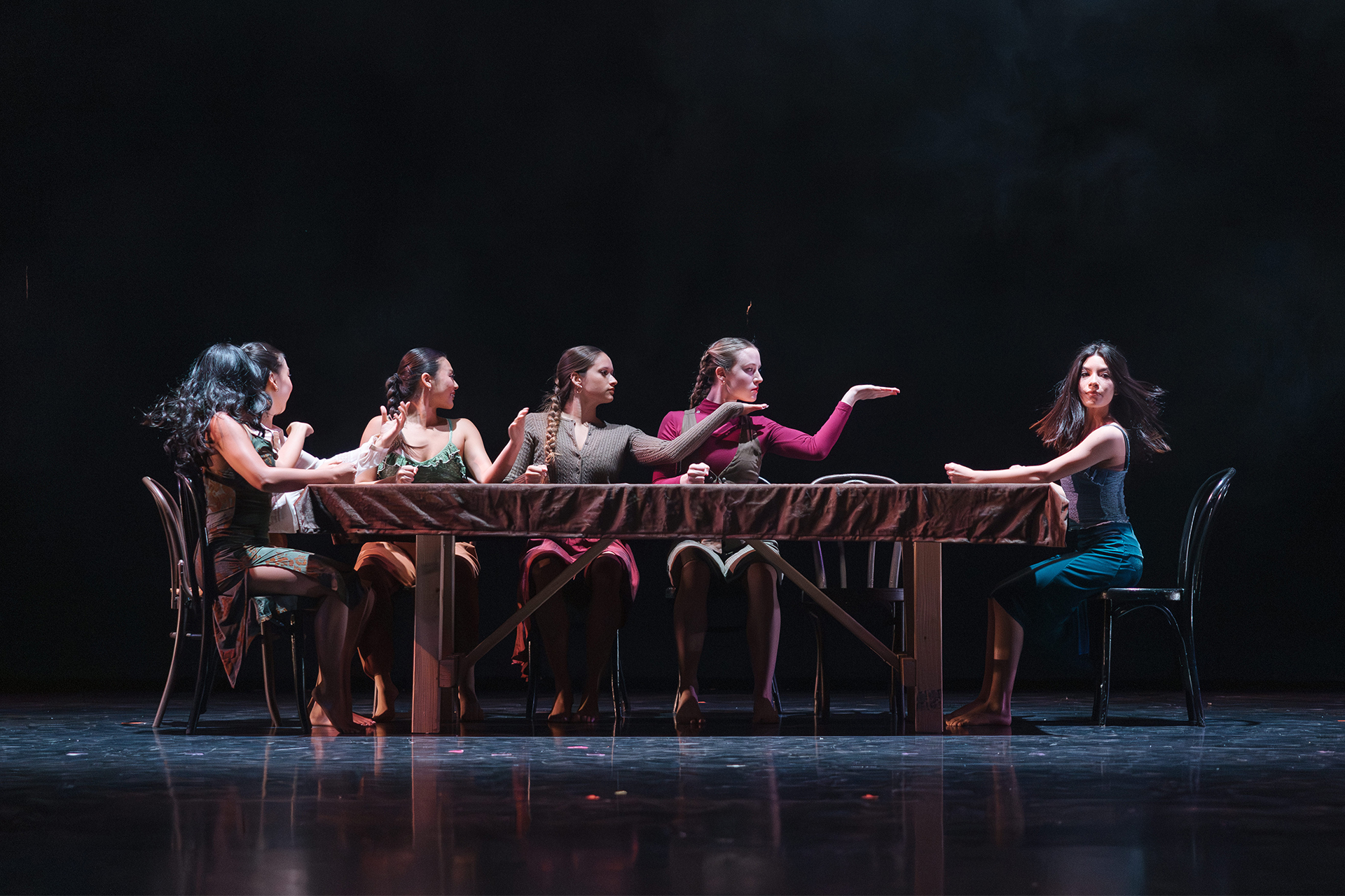 Dancers perform a theatrical piece seated along a draped table on a darkened stage, gesturing expressively toward one another.