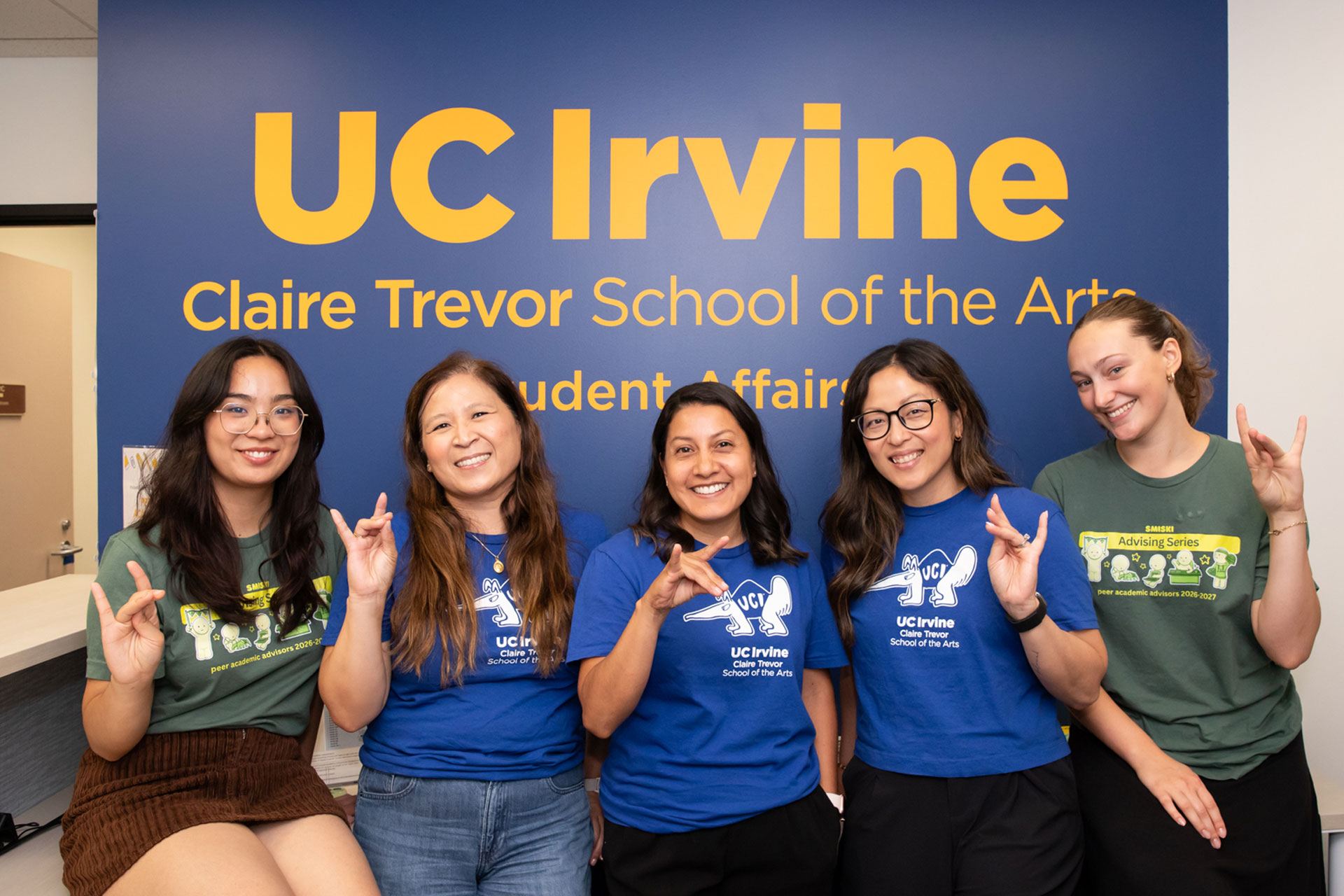 Five members of the UCI Claire Trevor School of the Arts Student Affairs team smile and make anteater hand signs in their office.