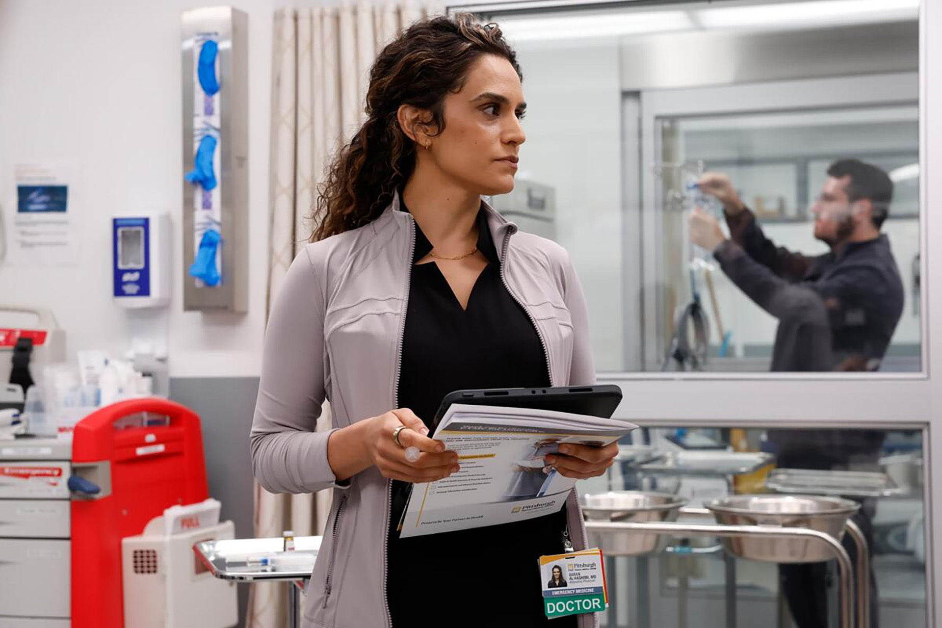 Sepideh Moafi with long curly dark hair stands in a hospital emergency room, wearing a black top under an open light gray zip-up jacket. She holds a tablet and printed paperwork while glancing off to the side with a focused expression. Her hospital ID badge reads "Baran Al-Hashimi, MD, Emergency Physician" with a green "DOCTOR" tag. In the blurred background, a man in dark scrubs adjusts an IV bag near a glass-walled trauma bay, with medical supply carts and glove dispensers visible in the foreground.