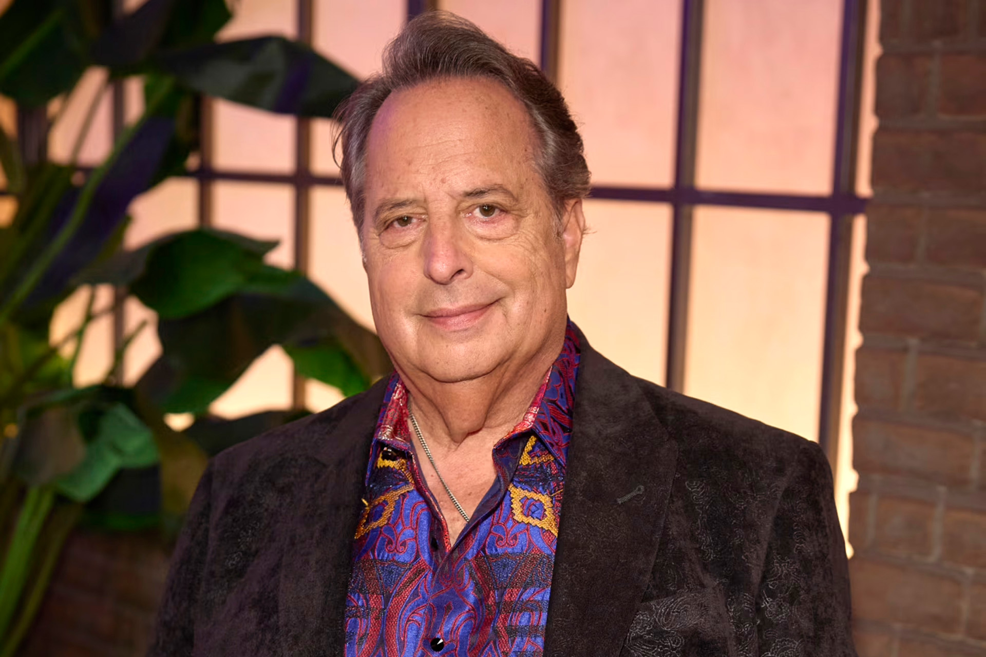 A close-up portrait of Jon Lovitz with graying hair, smiling slightly at the camera. He wears a dark textured blazer over a colorful blue, red, and gold patterned shirt with a silver chain necklace, standing in front of a paned window and greenery