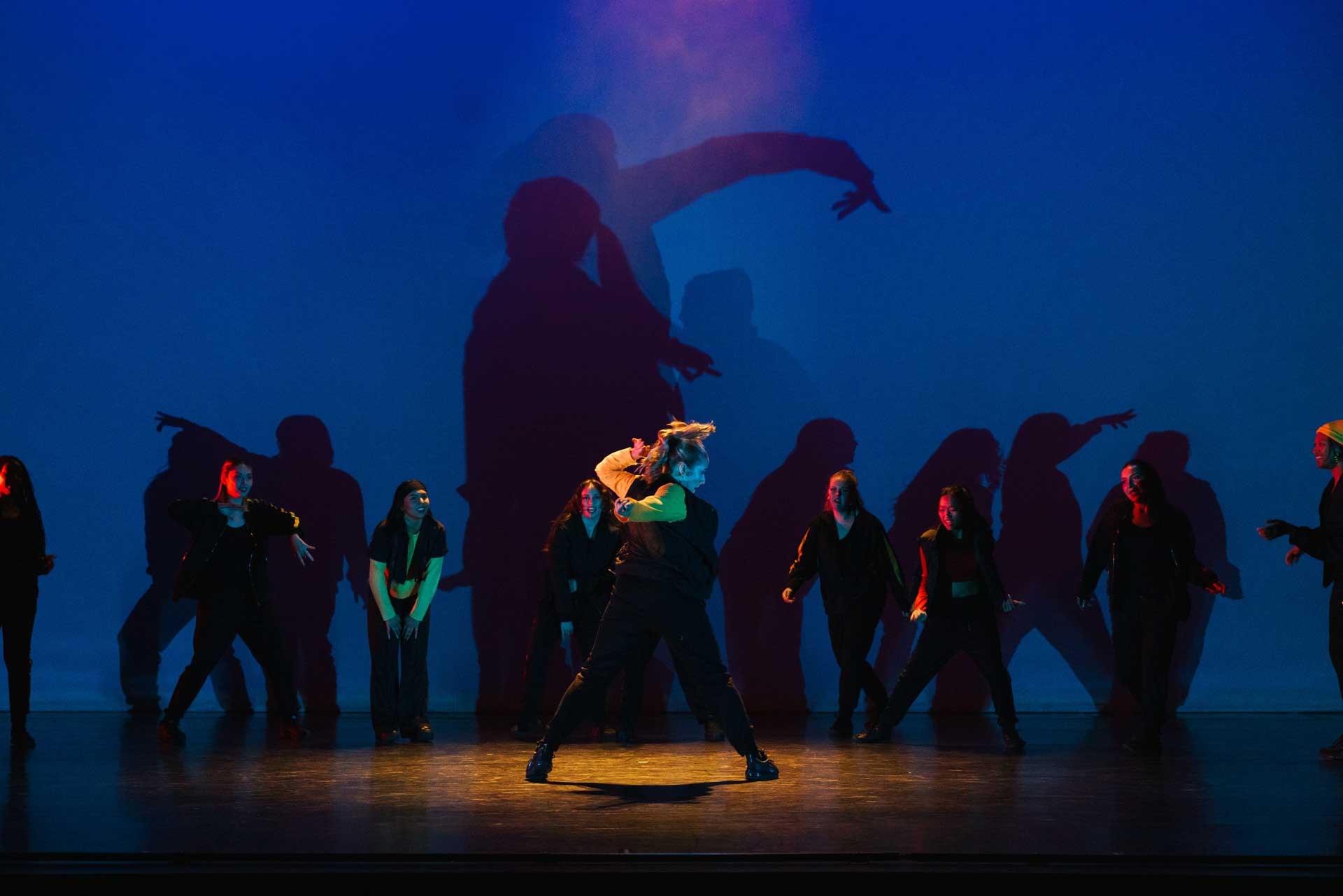 A group of dancers perform on stage under dramatic blue lighting, with bold shadows cast behind them that amplify movement and create a striking visual backdrop.