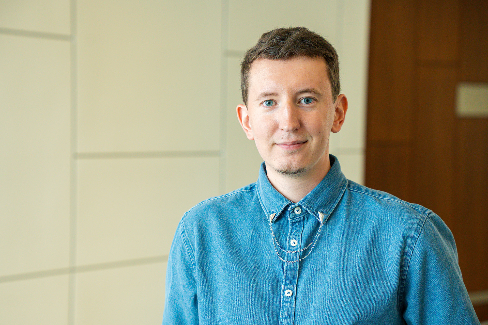 Portrait of a man with short brown hair and light eyes, wearing a denim button-up shirt with a chain collar accessory, smiling slightly at the camera in front of a light paneled wall.
