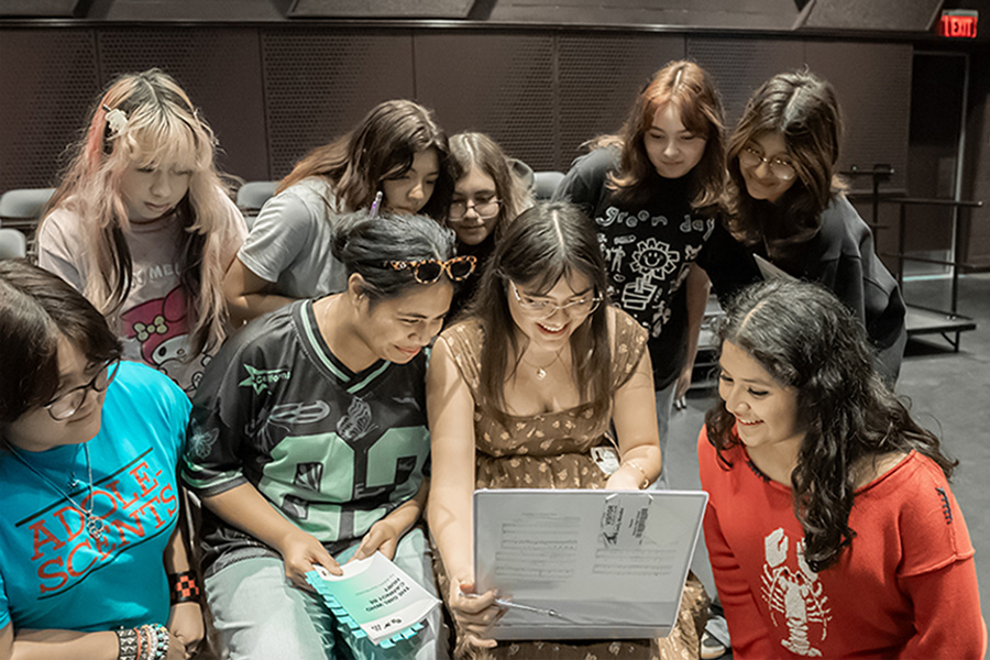 A group of students gather around a laptop, reviewing sheet music together backstage during a rehearsal.