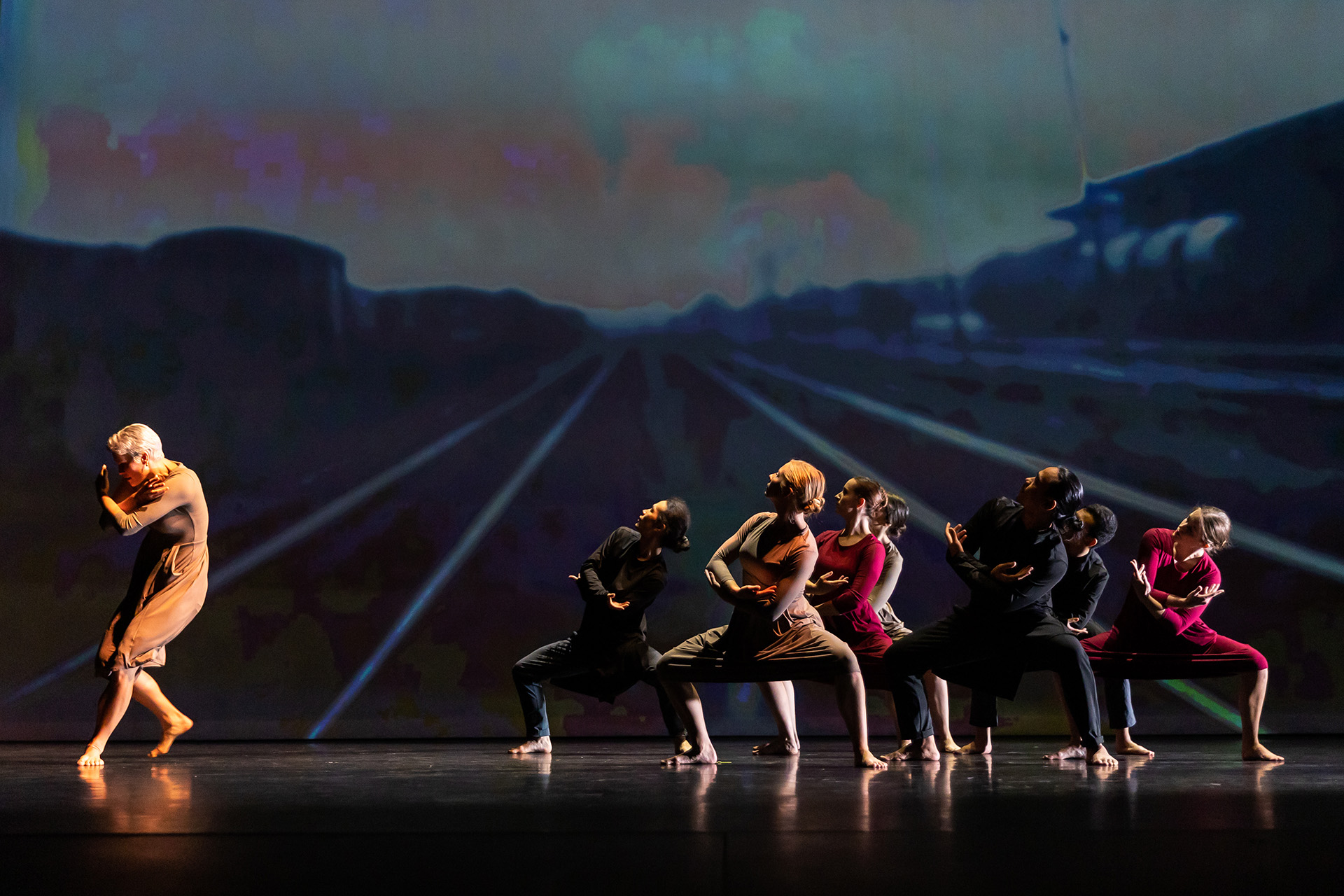 Boróka Nagy leads a line of dancers across a dimly lit stage, with a projected backdrop of train tracks behind them.