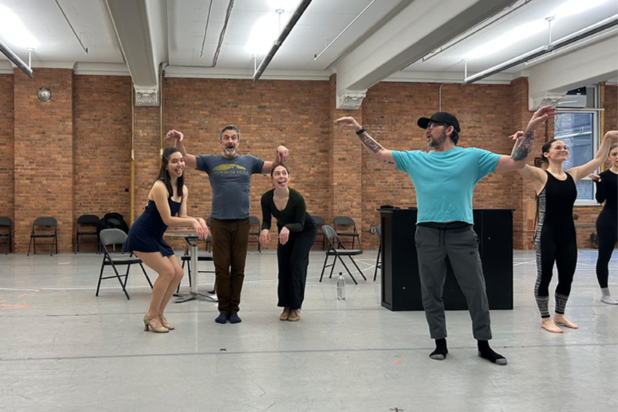 Andrew Palermo, in a teal shirt and black cap, leads dancers through choreography with arms outstretched in a brick-walled rehearsal studio.