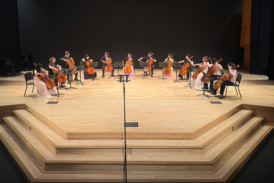 A cello ensemble performs in a semicircle on a wooden concert stage.