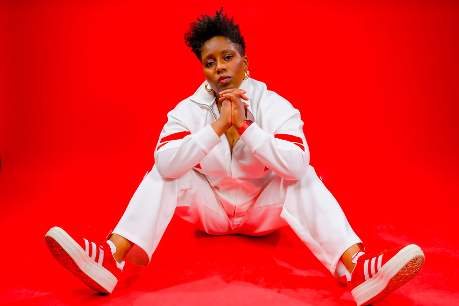 Cyrian Reed sits on the floor in a white tracksuit and red sneakers, chin resting on clasped hands, against a bright red backdrop.
