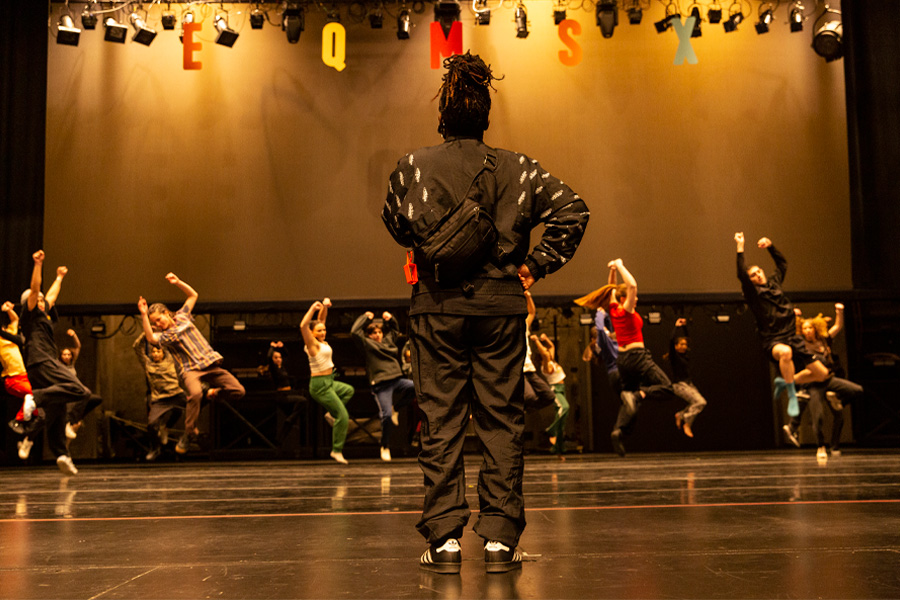 Cyrian Reed, seen from behind with hands on hips, watches students leap mid-air during a hip hop rehearsal onstage.