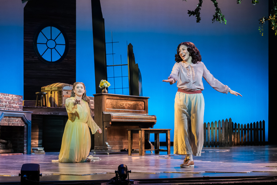 Jessica Lopez as Jo March, smiling with arms outstretched, crosses the stage toward a kneeling castmate in a yellow dress in UCI Drama's Little Women: The Broadway Musical.