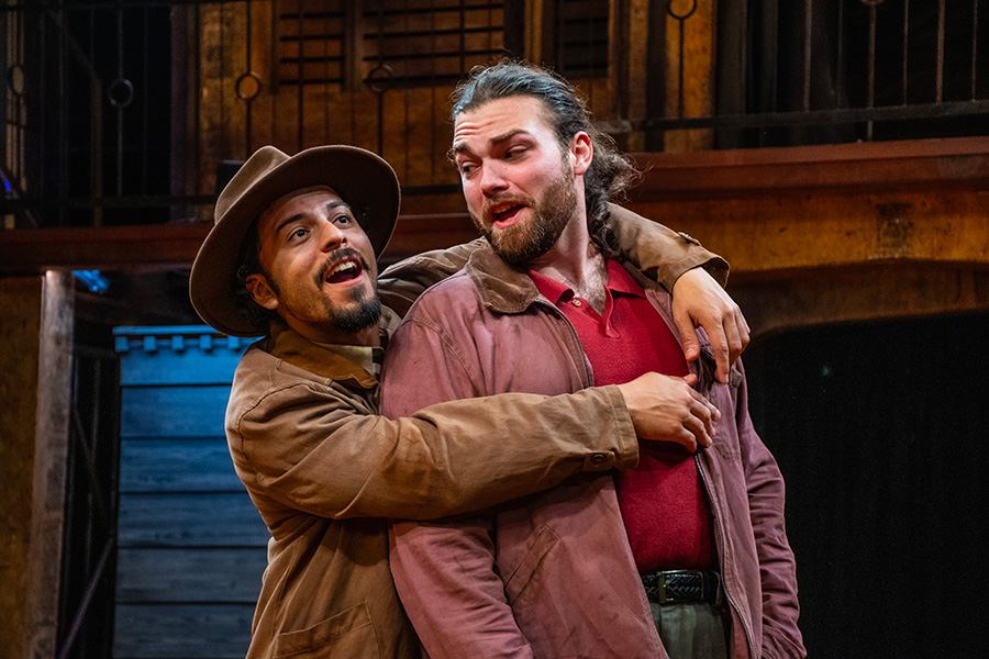 Heriberto Cruz Jr. as Mercutio, in a brown hat and jacket, laughs as he wraps his arms around a castmate from behind in New Swan Shakespeare Festival's Romeo & Juliet.