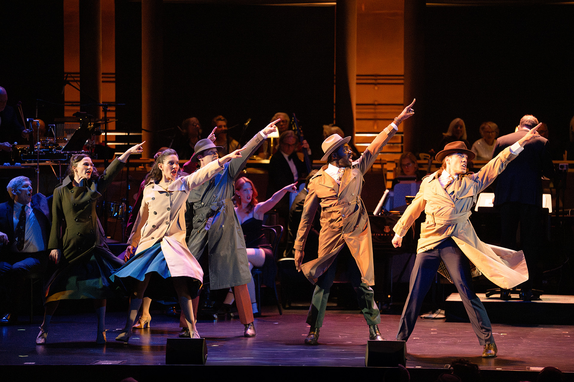 Performers in trench coats and fedoras point upward in unison onstage in a production choreographed by Andrew Palermo, with an orchestra and chorus seated behind them.