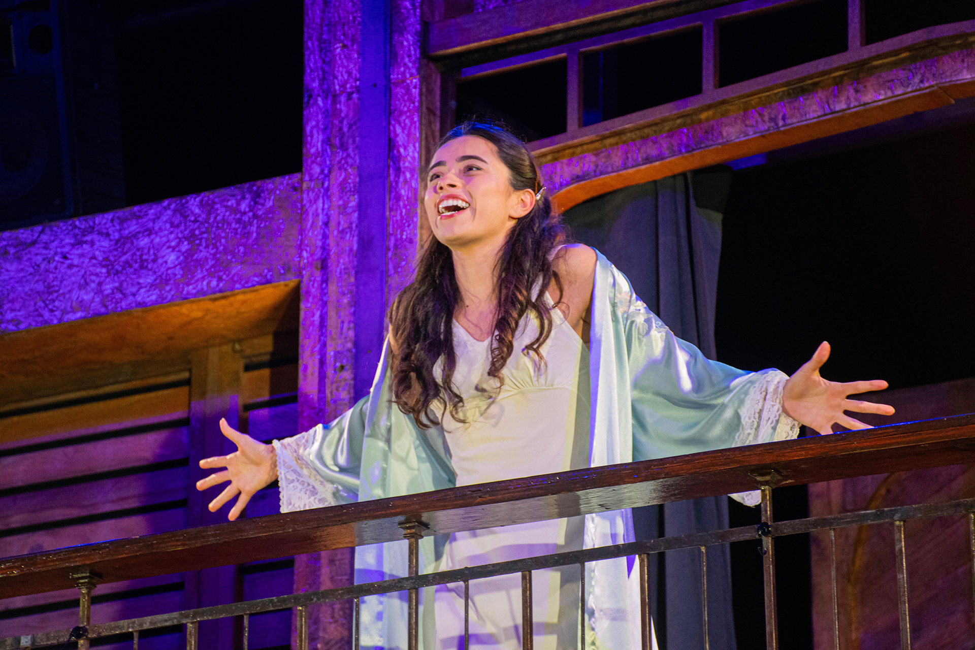 Lourdes Castillo as Juliet, arms outstretched and smiling on a balcony in a pale nightgown and robe, in New Swan Shakespeare Festival's Romeo & Juliet.