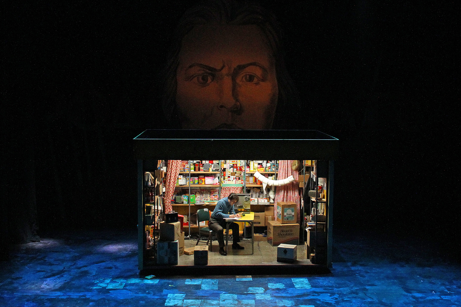 Production photo from Mother Russia, a small lit shop set cluttered with boxes and shelves of goods sits onstage as a man writes at a desk, beneath a giant painted face looming in the darkness above