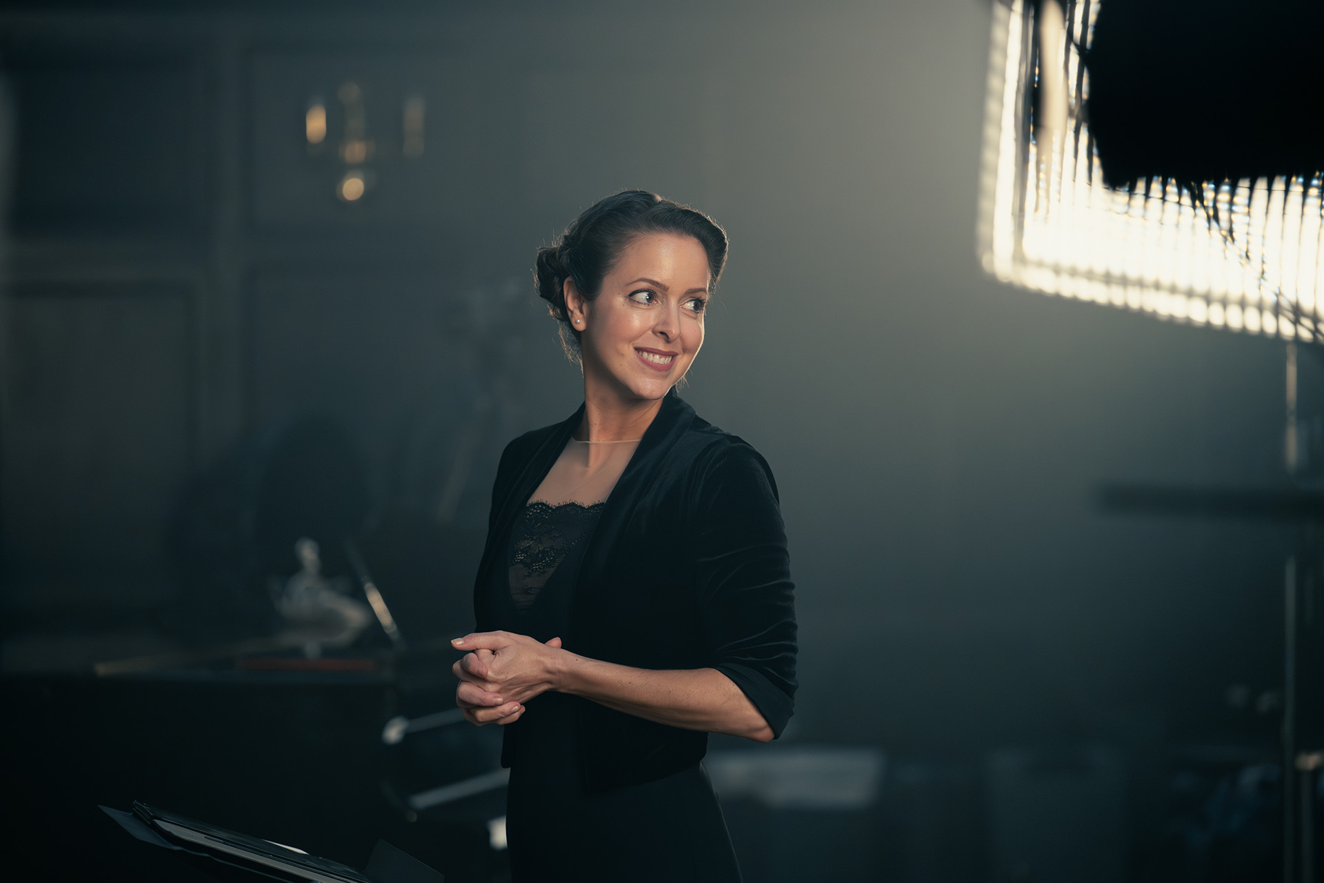 Irene Messoloras smiles and looks off to the side while standing with clasped hands, wearing a black velvet jacket over a lace top, during a portrait session with visible studio lighting in the background.