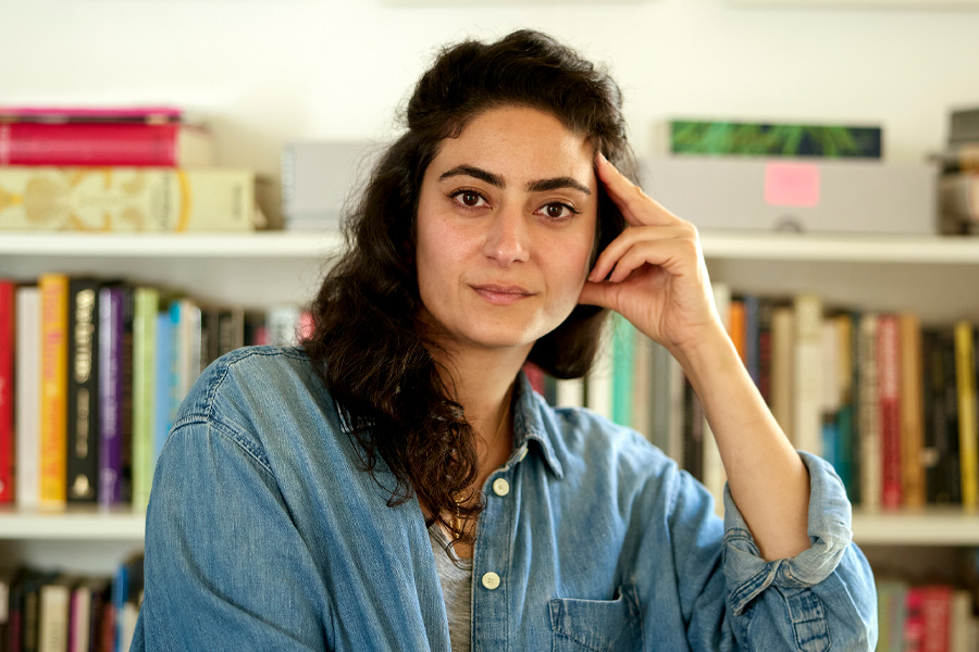 A woman with long dark curly hair, wearing a denim shirt, rests her temple against her fingertips and looks at the camera in front of a white bookshelf filled with colorful books.