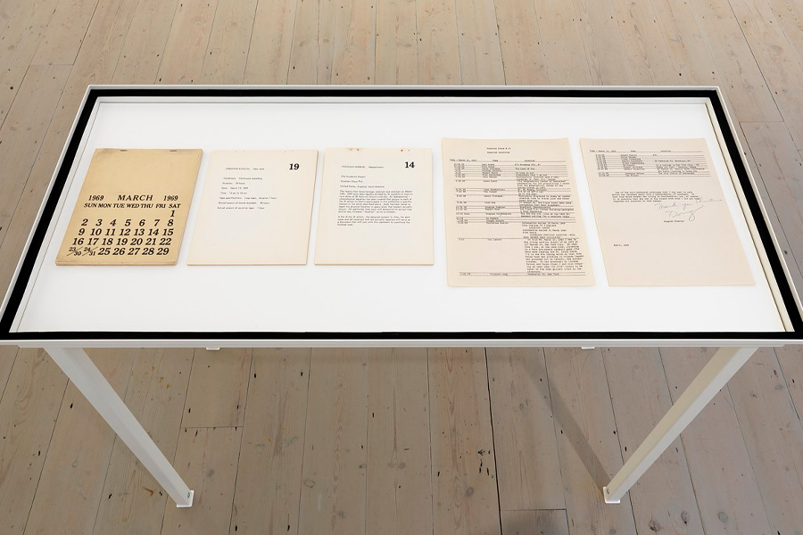A white display table in a gallery with wooden floors holds five archival documents under glass: a March 1969 calendar page, two typed artist pages for Christine Kozlov and Douglas Huebler, and two typewritten sheets listing times and locations, one signed.