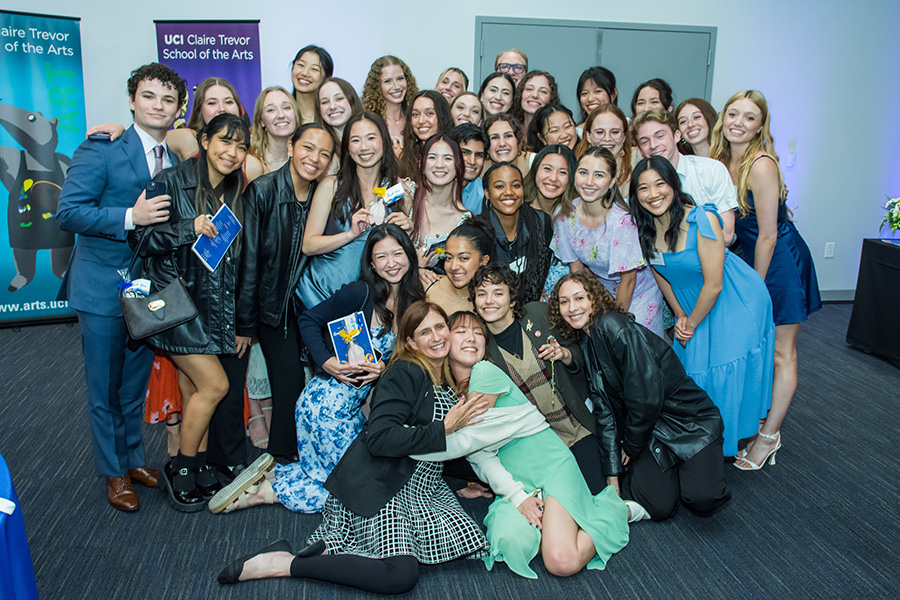 Dr. Kelli Sharp sits on the floor at the center of a large group of smiling UCI Dance students at the 2024 Annual Scholarship Dinner. Students gather closely around her, with several seated or kneeling in front and others standing behind.