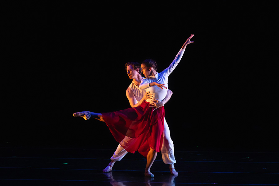 Two ballet dancers perform a duet on a dark stage. A woman in a white top and flowing red skirt extends one leg and reaches one arm high, while her partner, in white, supports her at the waist from behind.