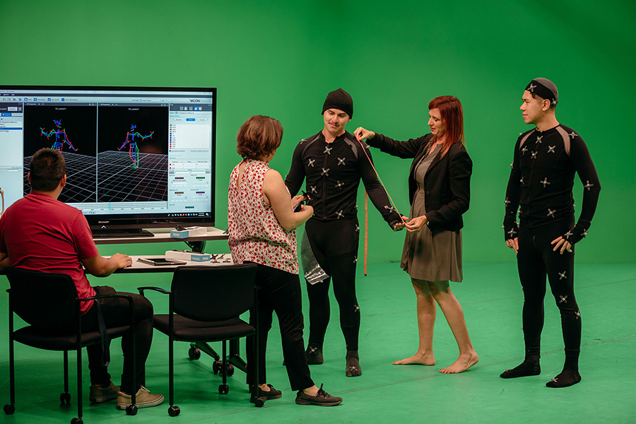 : Dr. Kelli Sharp works with an interdisciplinary research team in the Motion Capture Studio. Sharp, barefoot and holding a measuring tape, stands beside a student wearing a motion capture suit, while other team members observe and monitor movement data displayed on a screen.