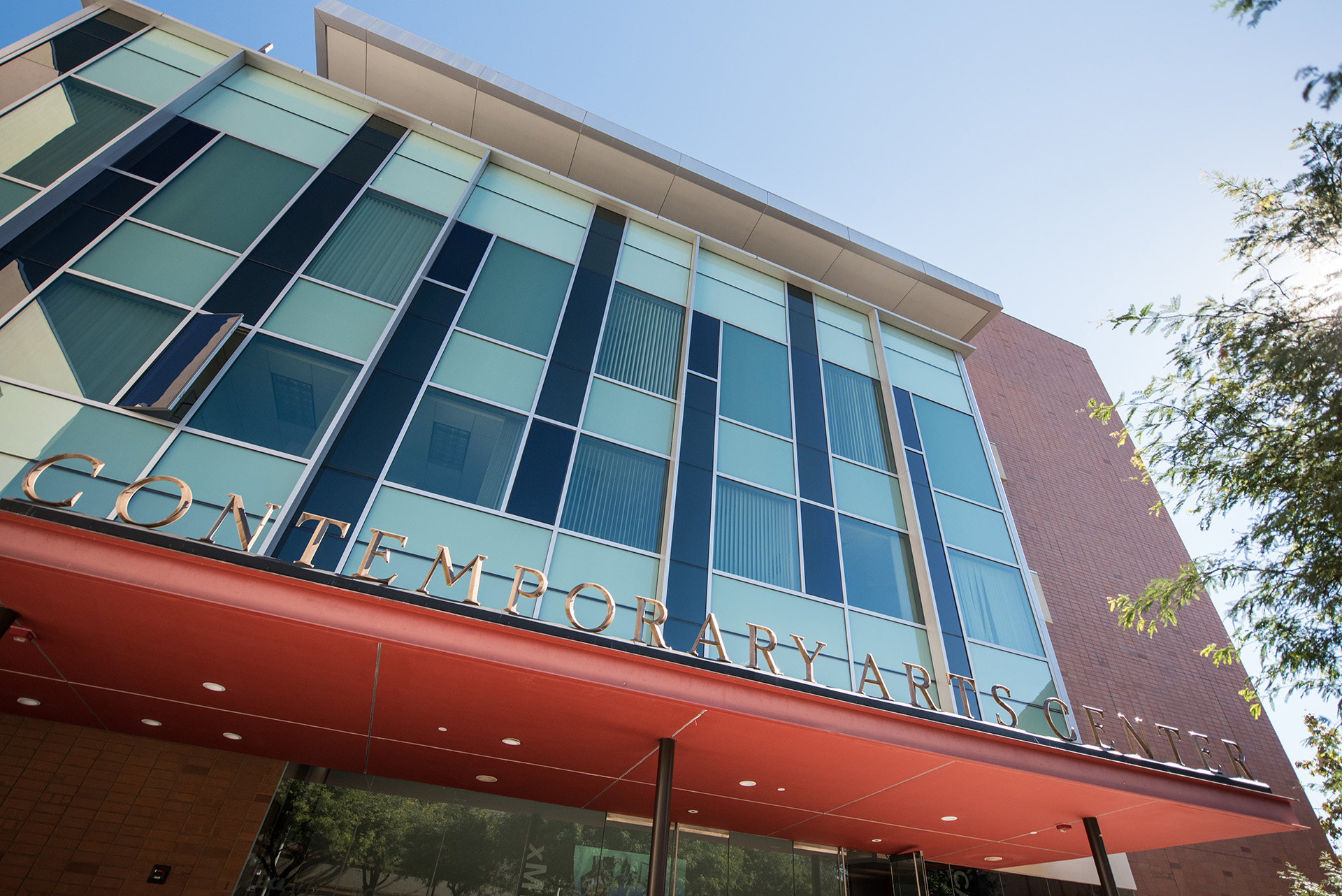 A low-angle view of the Contemporary Arts Center, with bronze letters spelling its name along a red-orange entrance canopy, a facade of teal and navy glass panels and a red brick wall against a clear blue sky.