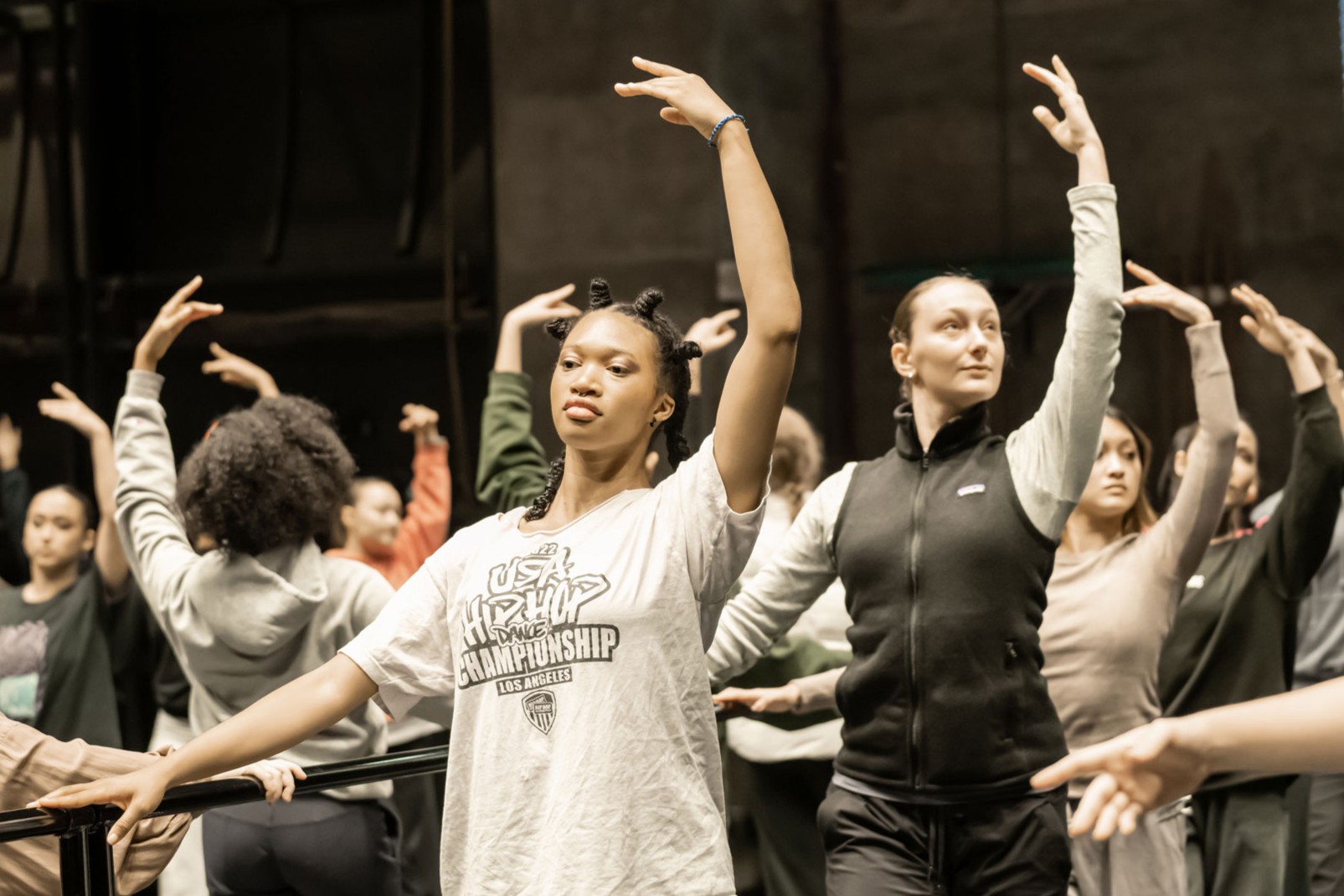 Dance students rehearse choreography in a studio, extending their arms in coordinated poses.