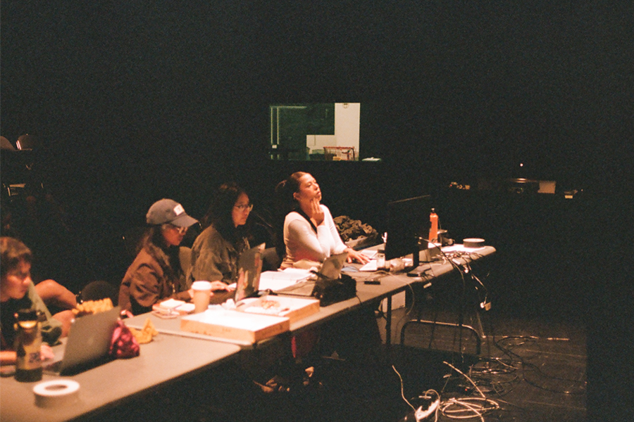 Members of the production team sit at a long table in a dark theater during rehearsal, working at laptops and monitors surrounded by scripts, cups and cables.