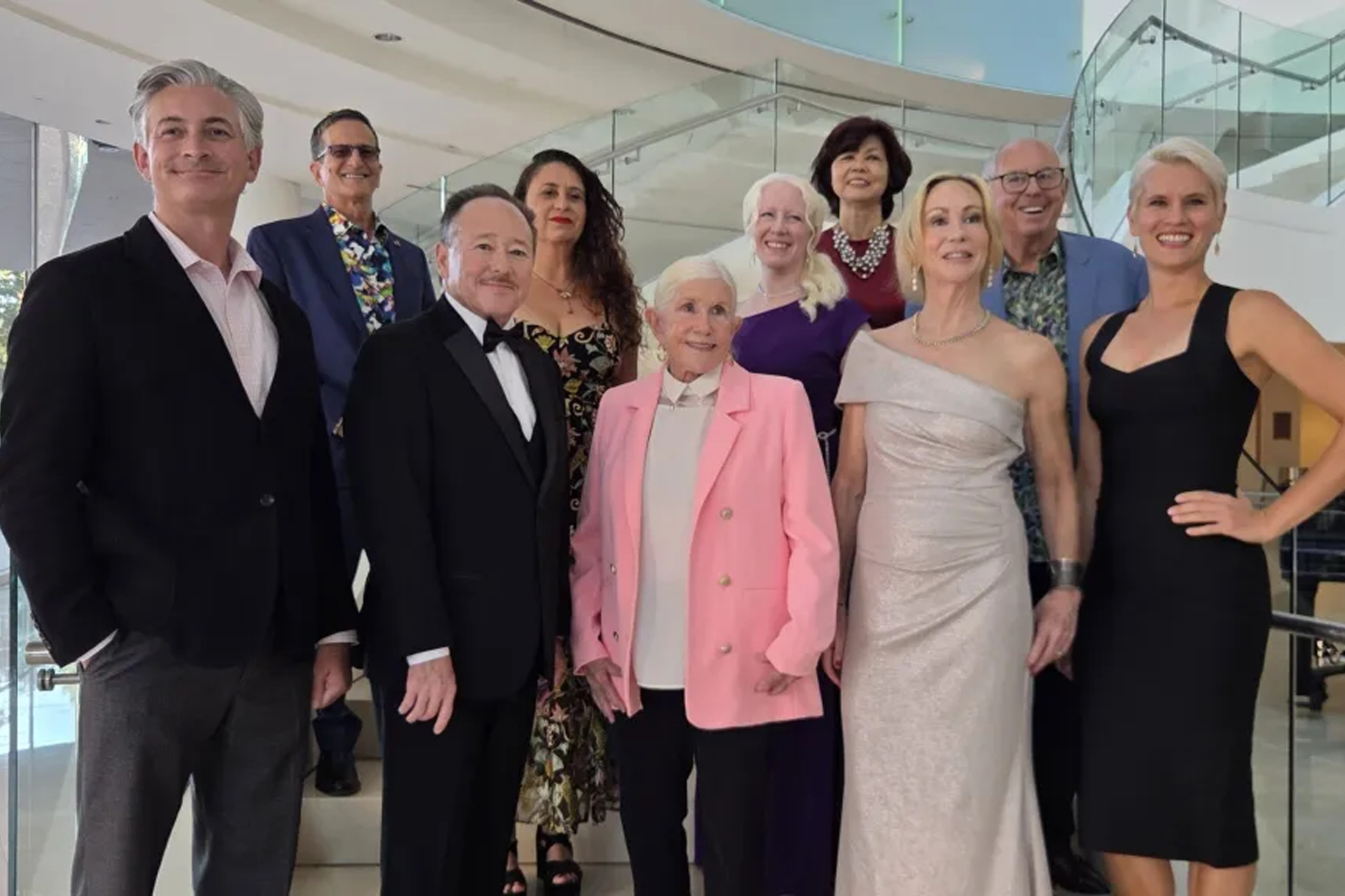 Nine OC Arts Awards honorees in formal attire pose together on a staircase with glass railings in a bright, modern lobby. A grand piano is visible behind them.