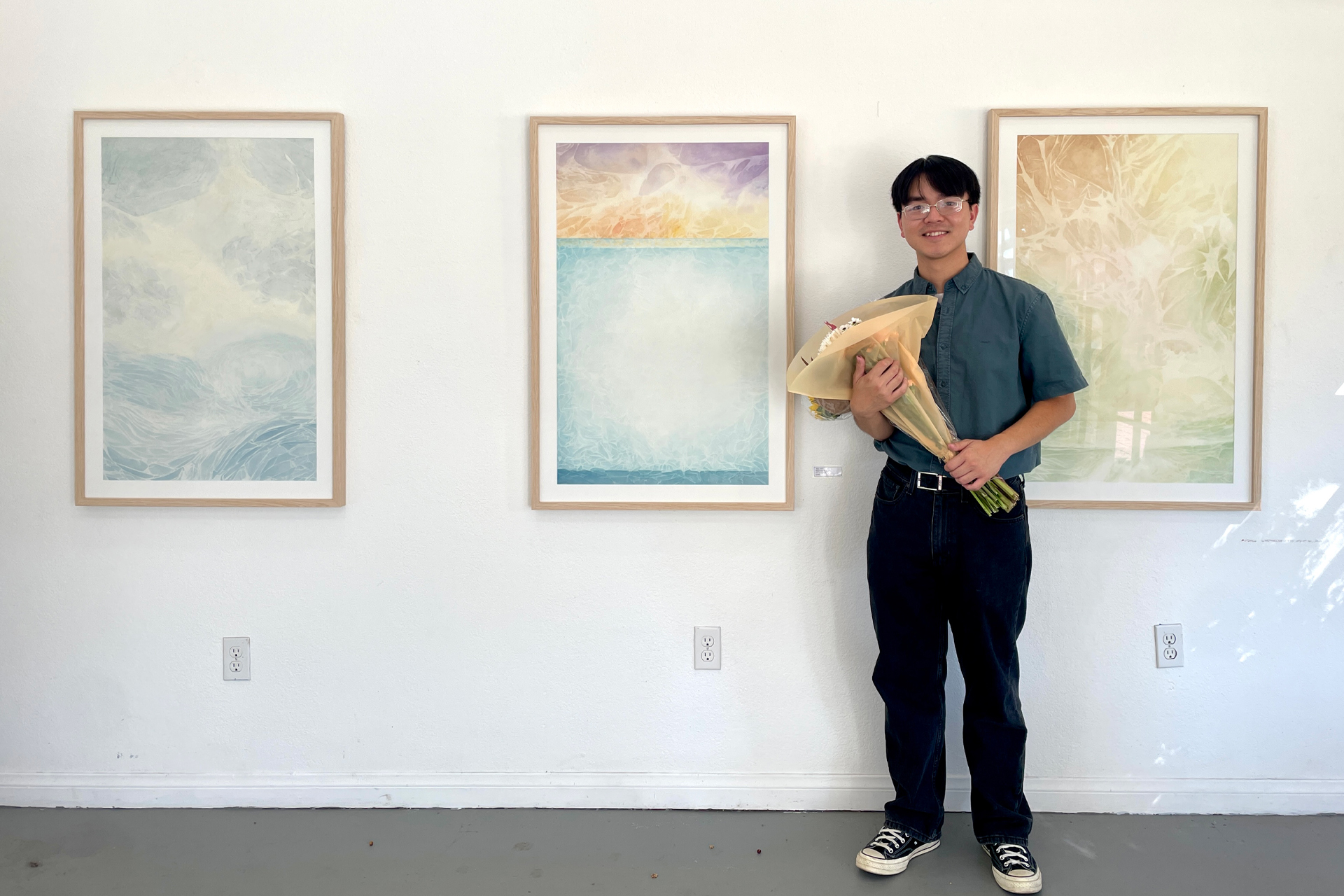 An artist holding a bouquet of flowers smiles beside three framed works on a white gallery wall. The works are soft, layered abstractions in blue, gold and green that suggest water and sky.