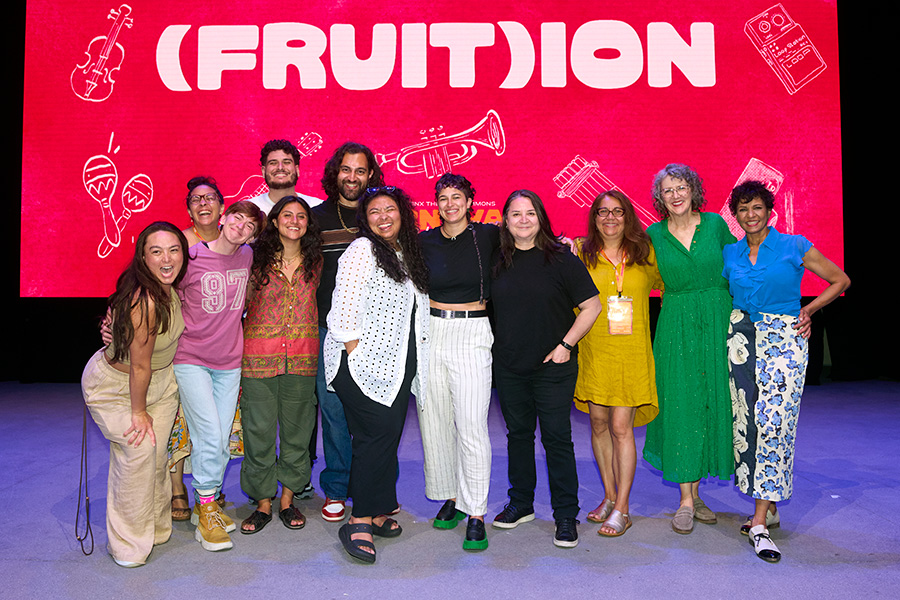 uliette Carrillo, in a yellow dress, poses with 10 smiling cast and creative team members on a stage in front of a large red screen reading “(Fruit)ion,” decorated with white line drawings of musical instruments.