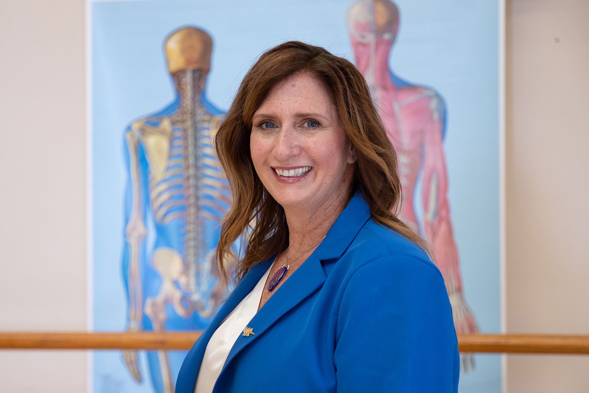 Dr. Kelli Sharp smiles at the camera, wearing a bright blue blazer over a white blouse. Anatomical illustrations of the human skeletal and muscular systems are displayed on the wall behind her.