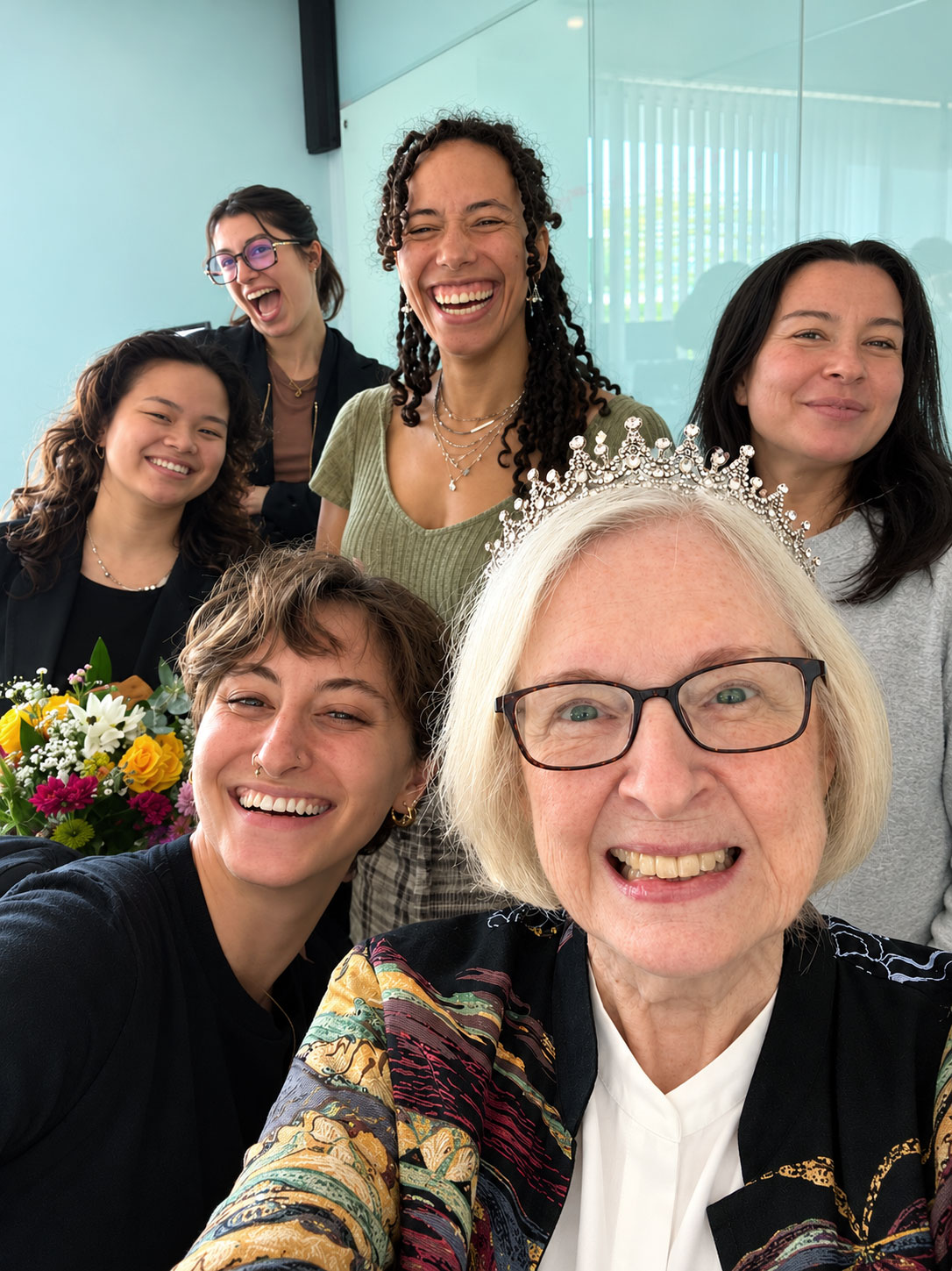 Professor Jennifer Fisher and students. Six women smile and laugh together in a group selfie, with one wearing a silver tiara and another holding a colorful bouquet of flowers.