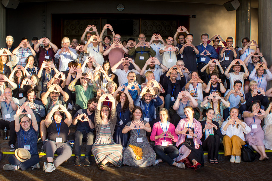 A large group of conference attendees wearing lanyards pose together on steps in a sunlit lobby, each making a triangle shape with their hands.