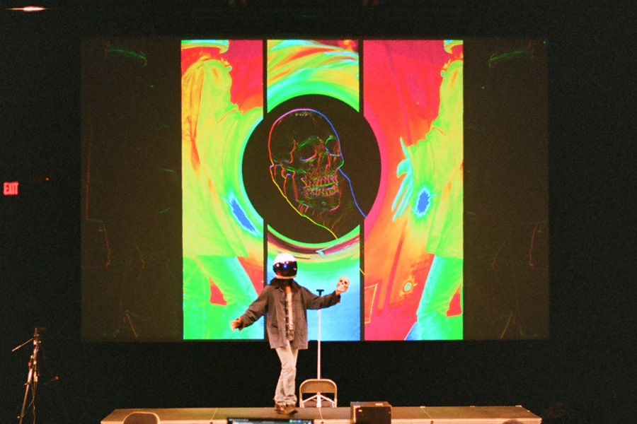 Kurt Kana in a white helmet stands on stage holding up a small skull during a rehearsal of No-No Boy, a Solo Show. Behind them, a large projection shows a skull in neon green, red and blue.