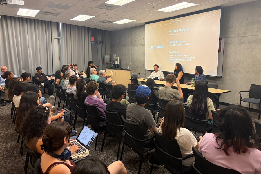 A packed audience sits in rows facing three panelists at a table at the front of a classroom. A projected slide behind them reads “Our Panel” and lists the speakers’ names and titles. Several audience members take notes, one on a laptop.