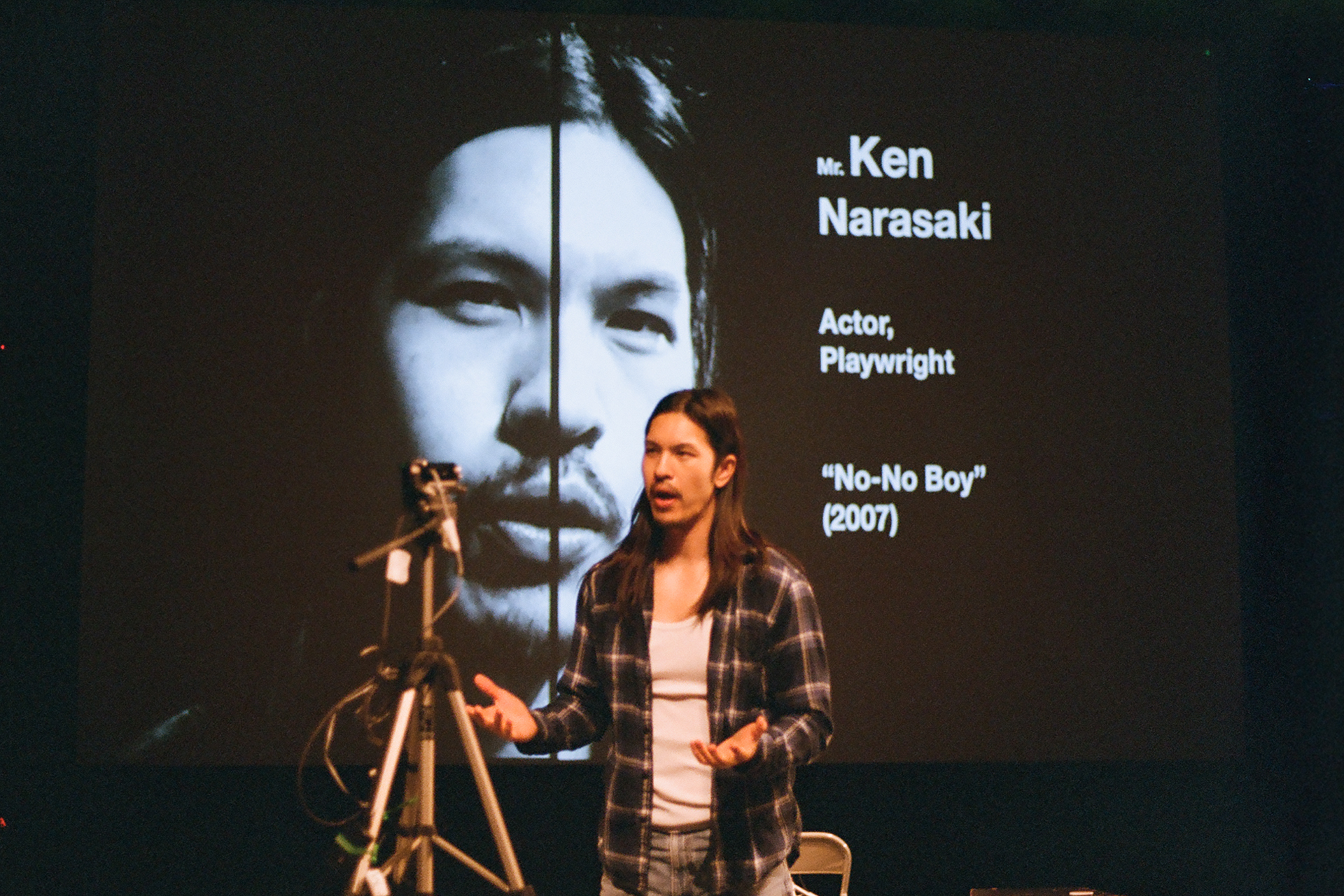 Kurt Kana, wearing a plaid shirt over a white tank top, speaks on stage with his hands open beside a camera on a tripod. A projection behind him shows a black-and-white close-up of a face and the words "Mr. Ken Narasaki, Actor, Playwright, 'No-No Boy' (2007)."
