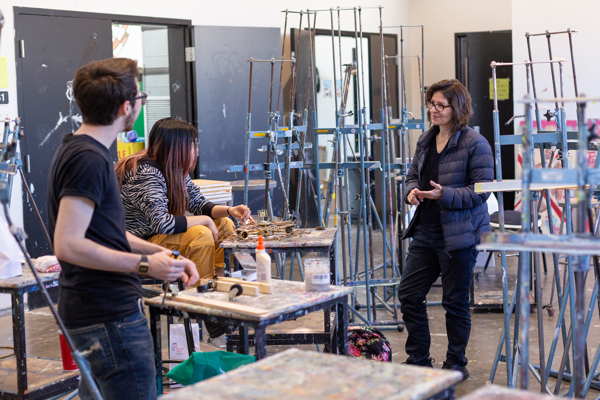 Professor Monica Majoli talks with two students in a paint-spattered studio filled with metal easels. One student sits at a worktable assembling a small brass and metal sculpture, while the other stands nearby holding a tool.