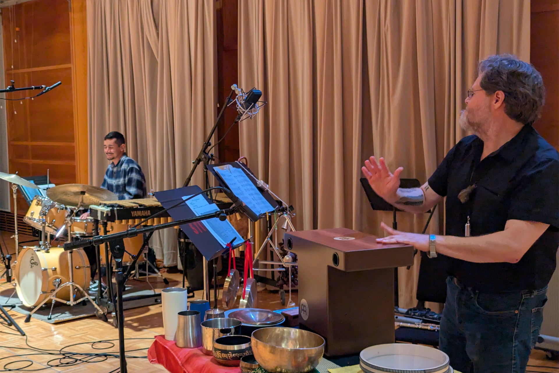 Camilo Zamudio plays marimba behind a drum kit as co-leader Wilfrido Terrazas gestures beside a cajón and a table of singing bowls and frame drums during a rehearsal in a wood-paneled studio.