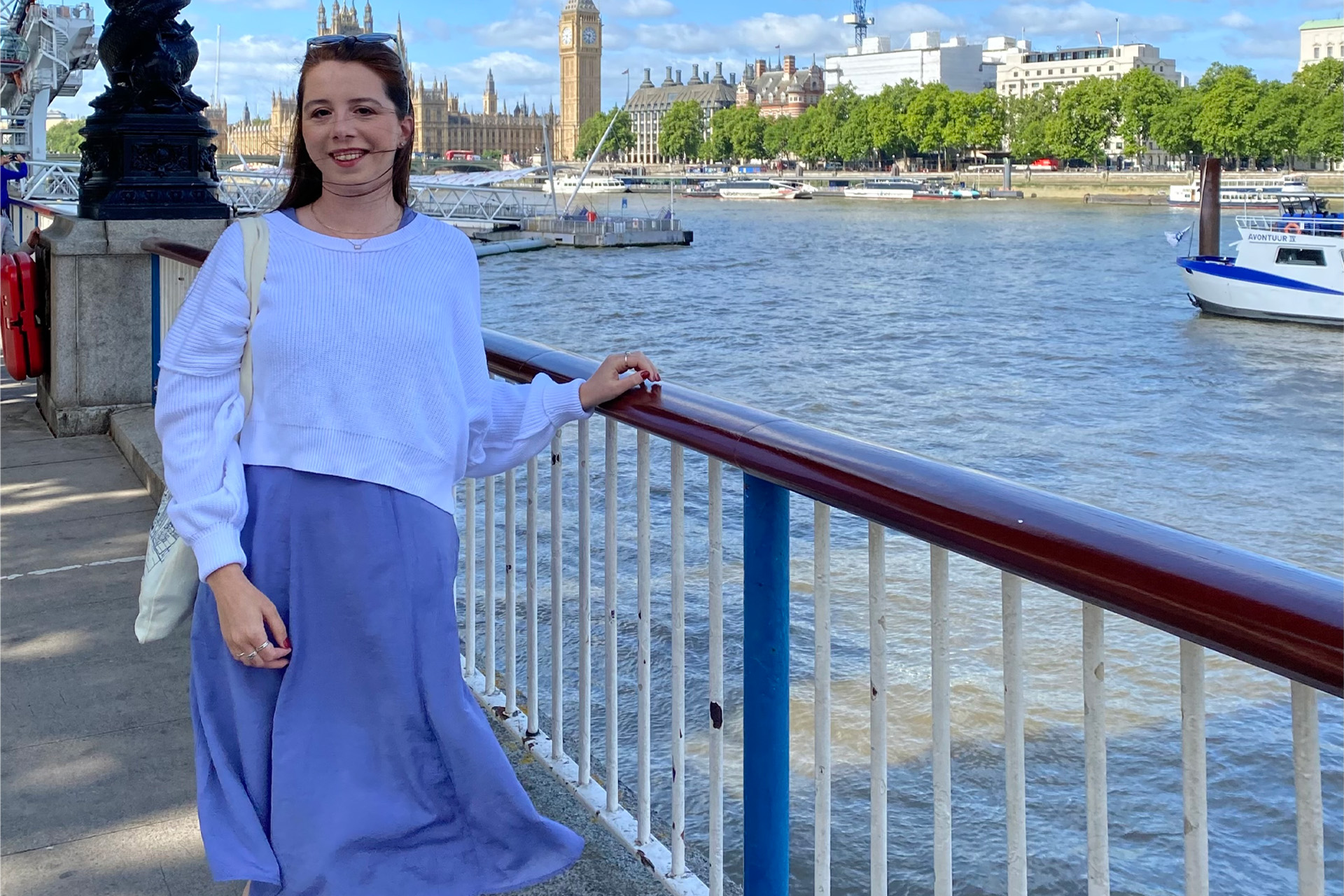 Emily Parise, in a white sweater and periwinkle dress, smiles beside a railing on the River Thames, with Big Ben and the Houses of Parliament in the background.