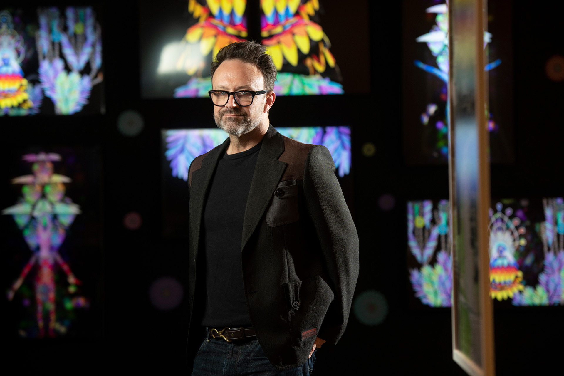 Matt Bailey, wearing a dark blazer over a black T-shirt and black-framed glasses, stands with one hand in his pocket in a darkened gallery surrounded by glowing, symmetrical digital artworks in vivid rainbow colors.