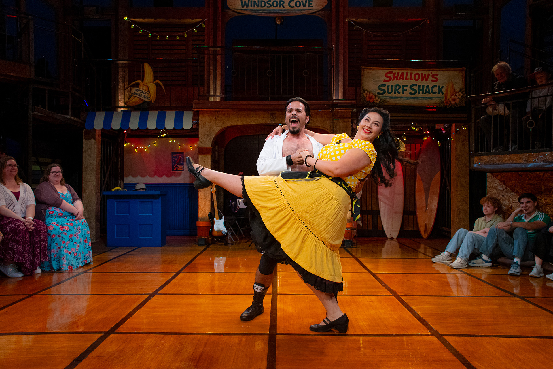 On a sunlit wooden stage dressed as a beachside boardwalk, with signs for “Windsor Cove” and “Shallow’s Surf Shack,” a laughing man in an open white shirt dips a smiling woman in a yellow polka-dot top and full yellow skirt as she kicks one leg high. Audience members seated around the stage edge look on and smile.